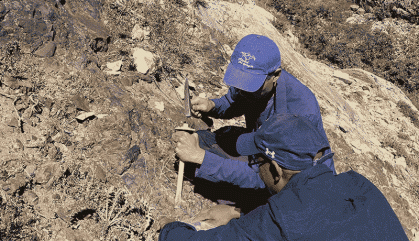 Two people crouch to the ground near a mountain and collect rock samples.