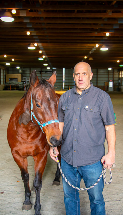 Man in blue shirt and blue jeans stands next to a therapy horse.